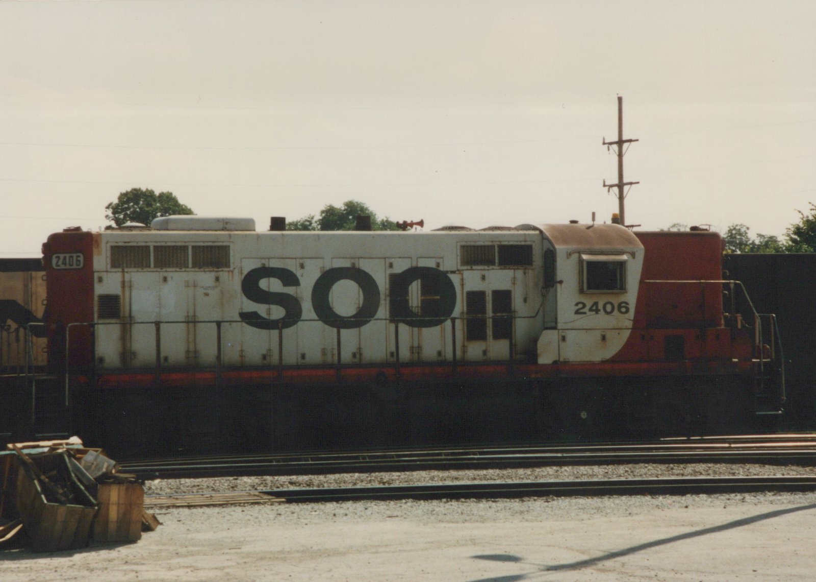 Side view of a white and red Soo Line diesel locomotive numbered 2406, parked in a rail yard with multiple tracks and trees in the background.
