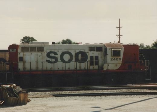 Side view of a white and red Soo Line diesel locomotive numbered 2406, parked in a rail yard with multiple tracks and trees in the background.