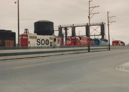 A line of diesel locomotives from Soo Line, Canadian Pacific, and GATX sits on a bridge over a road, with a water tower and industrial structures in the background.