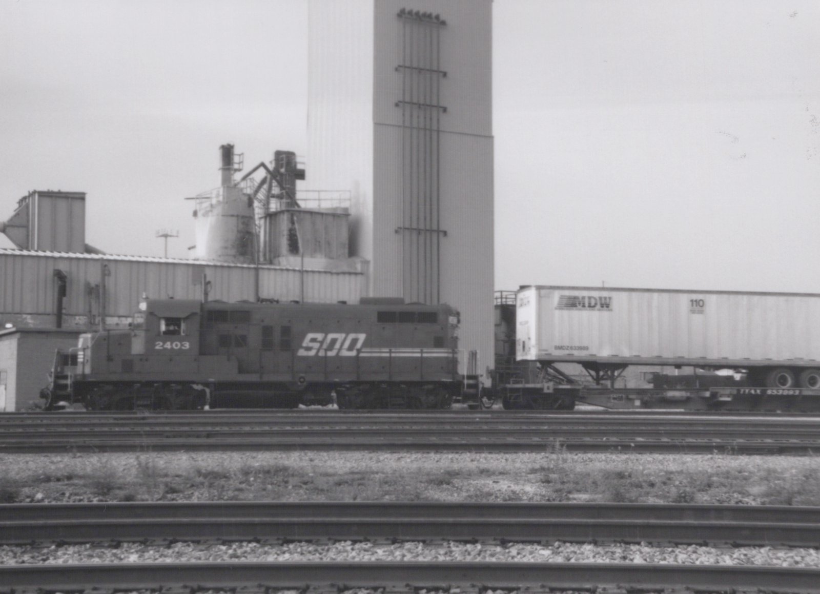 A black and white photo of a SOO Line diesel locomotive numbered 2403, pulling a freight car with a trailer, set against an industrial backdrop.