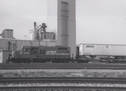 A black and white photo of a SOO Line diesel locomotive numbered 2403, pulling a freight car with a trailer, set against an industrial backdrop.