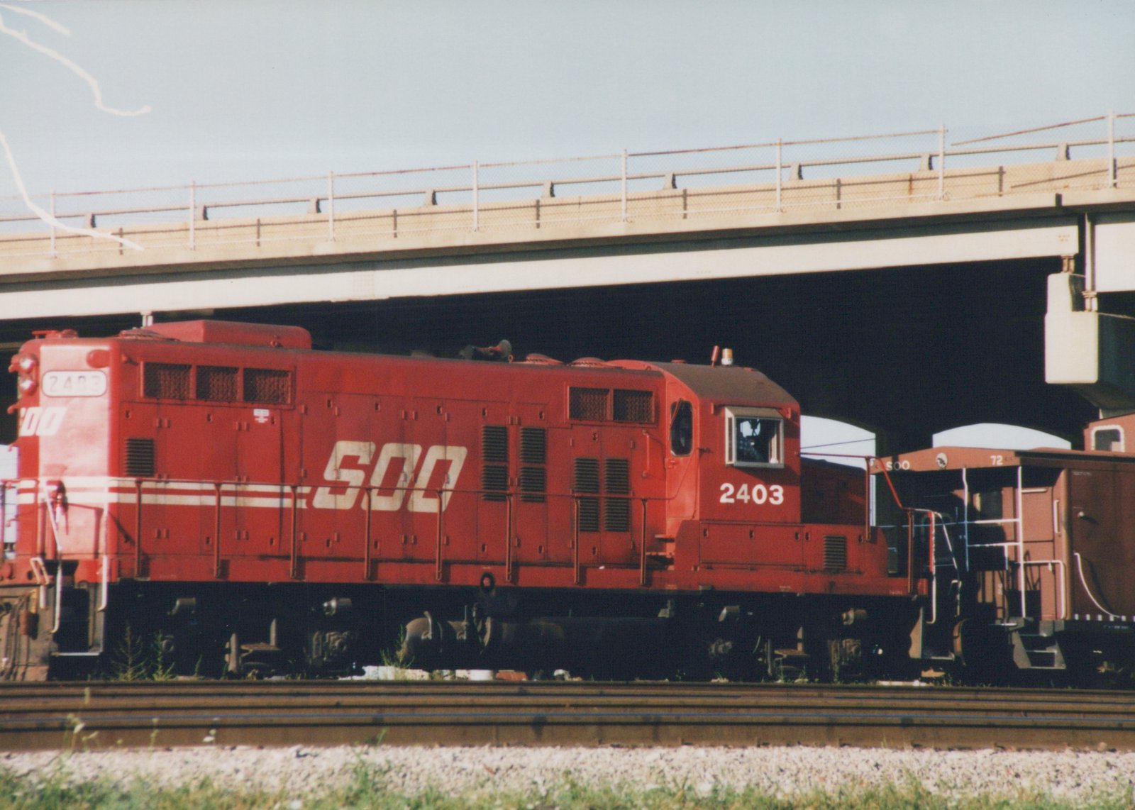A red Soo Line diesel locomotive number 2403 and a red caboose are on railroad tracks beneath a concrete highway bridge on a clear day.