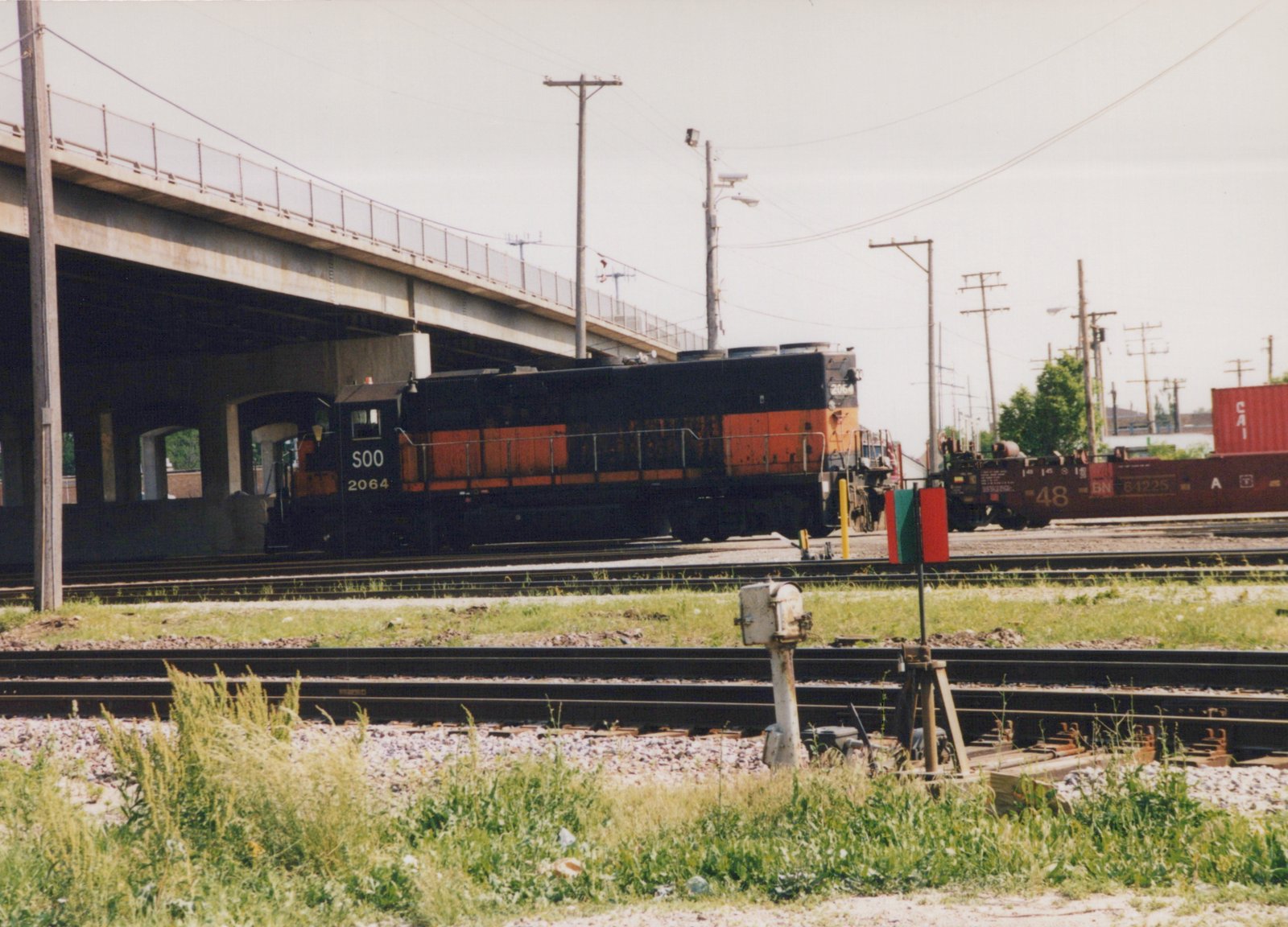A black and orange Soo Line diesel locomotive numbered 2064 sits on tracks under an overpass, with a dark brown Burlington Northern freight car nearby in a grassy yard.