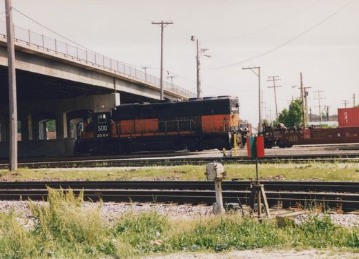 A black and orange Soo Line diesel locomotive numbered 2064 sits on tracks under an overpass, with a dark brown Burlington Northern freight car nearby in a grassy yard.