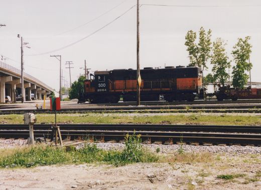A black and orange SOO Line diesel locomotive numbered 2064 is parked on railroad tracks beside an elevated highway, with a red freight car marked BN 48 behind it.