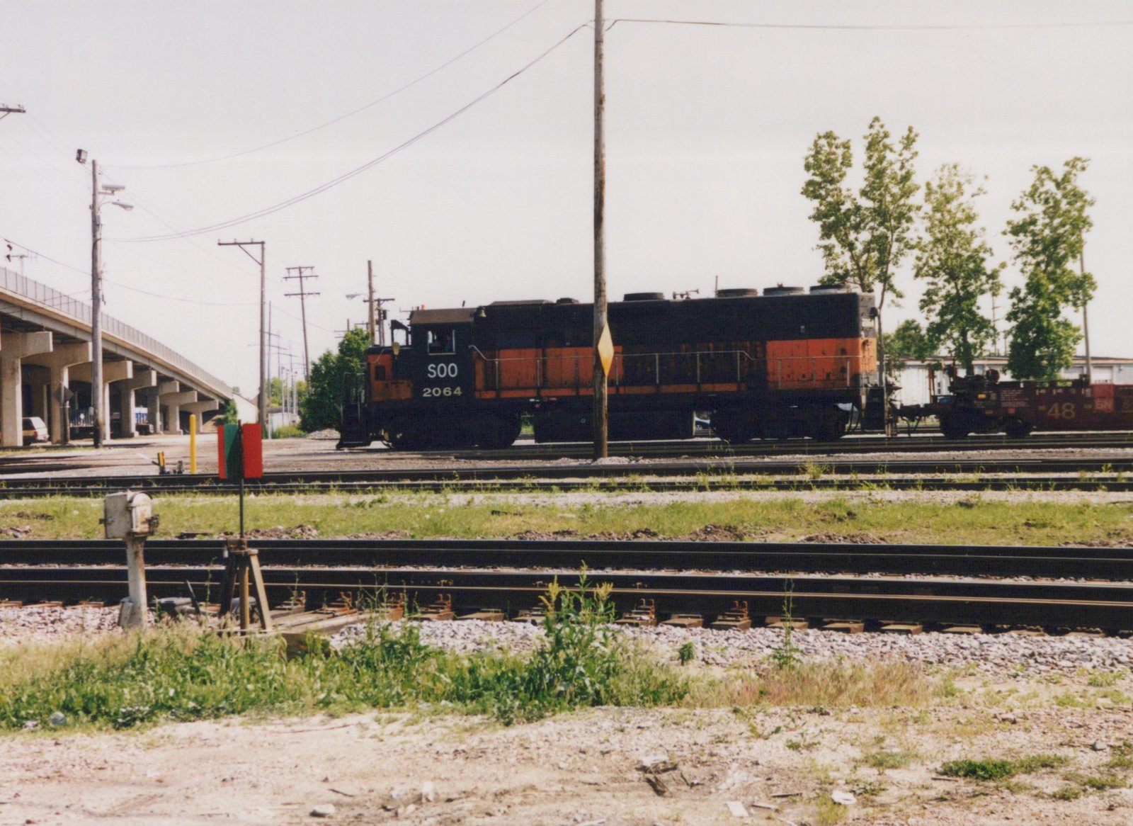 A black and orange SOO Line diesel locomotive numbered 2064 is parked on railroad tracks beside an elevated highway, with a red freight car marked BN 48 behind it.