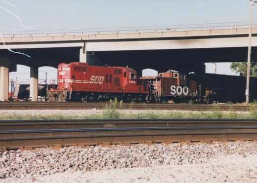 A red Soo Line diesel locomotive numbered 2403 and a matching caboose numbered 72 travel under an overpass on a sunny day in Franklin Park, Illinois.