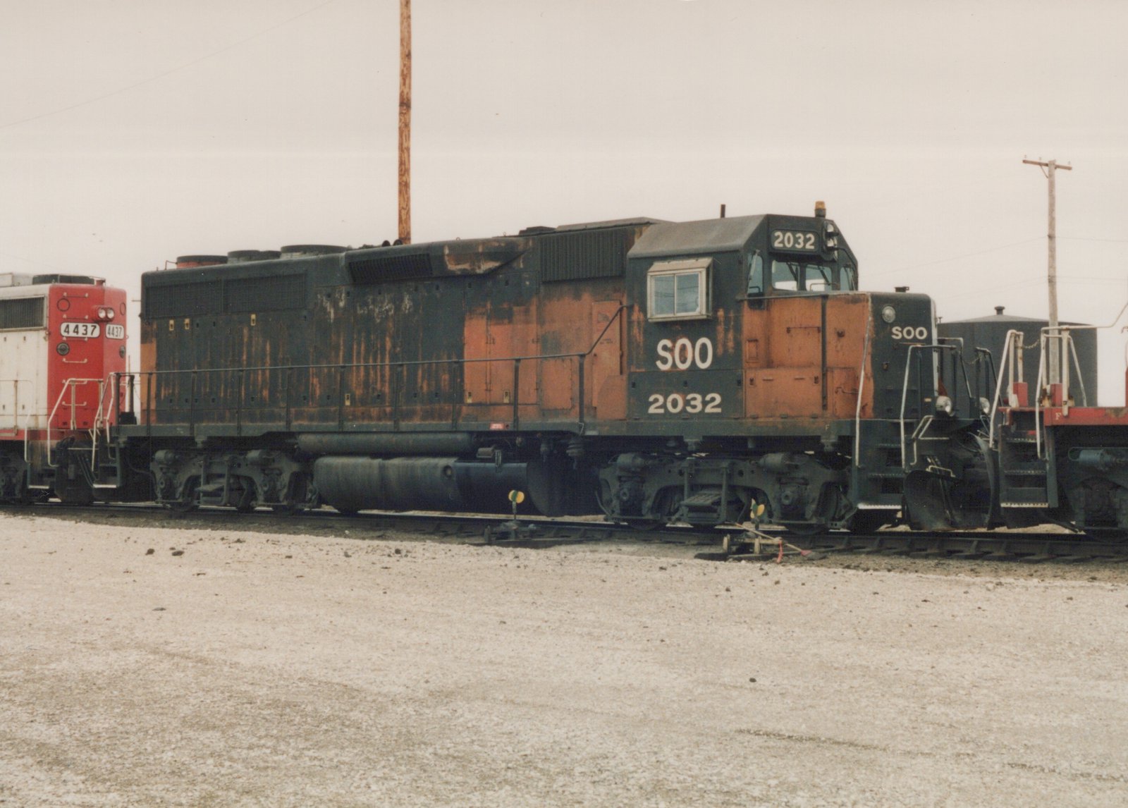 A weathered black and orange Soo Line diesel locomotive, number 2032, is pictured from the side on a gravel yard track, with a red and white Soo Line locomotive, number 4437, partially visible behind it.