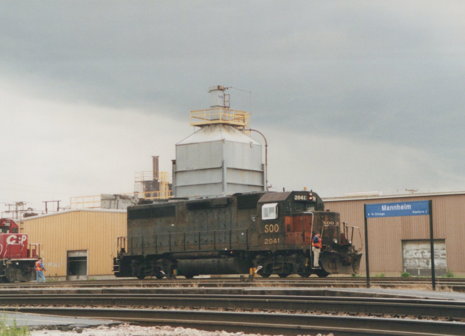 A black SOO Line diesel locomotive, number 2041, is stationary on tracks at the Mannheim station yard, with a person standing on its steps and another person on the ground nearby; a red C:P locomotive is visible in the background.