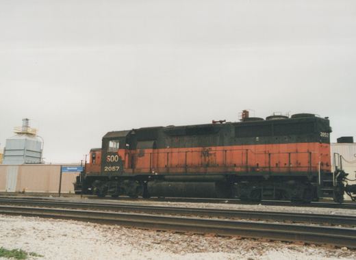 A side view of SOO Line diesel locomotive 2057, painted orange and black, on railroad tracks with an industrial building and a 'Mannheim' station sign in the background under an overcast sky.