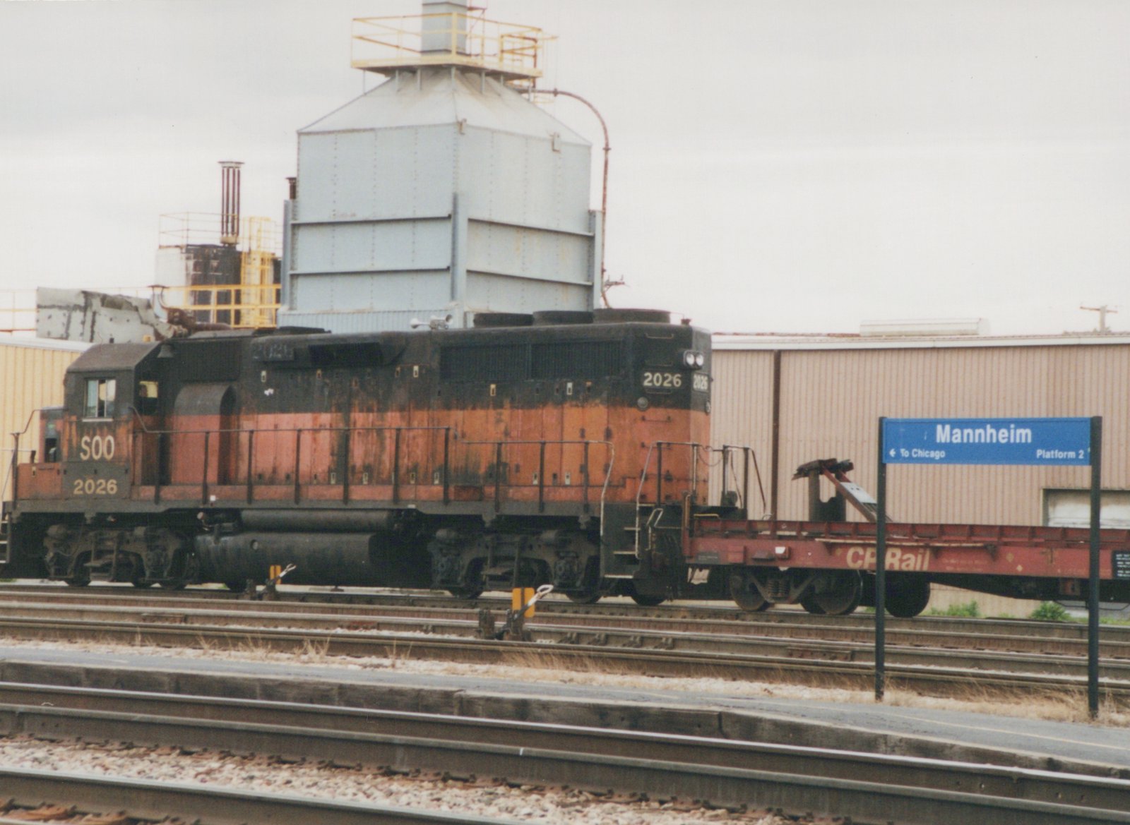 Side view of a Soo Line locomotive numbered 2026 and a CP Rail flatcar at Mannheim station, with a blue platform sign indicating 'To Chicago'.