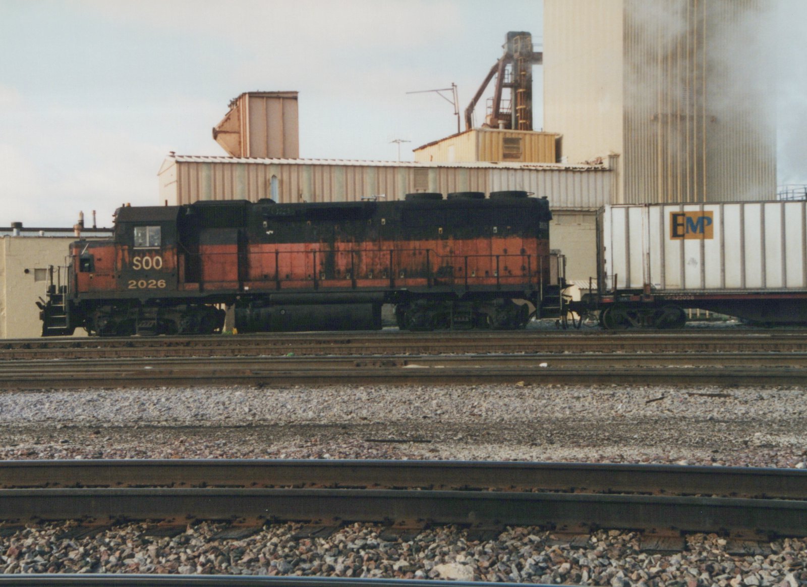 A side view of a SOO Line diesel locomotive numbered 2026, painted orange and black, coupled to a flatcar hauling a white trailer with 'EMP' on its side, set against an industrial backdrop with grain silos.