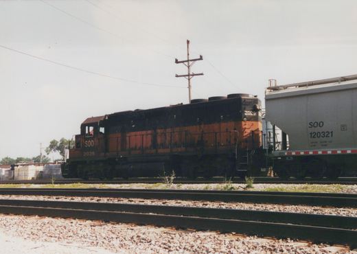 A black and orange Soo Line diesel locomotive numbered 2026 is pulling a freight car along railroad tracks with overhead power lines in the background.