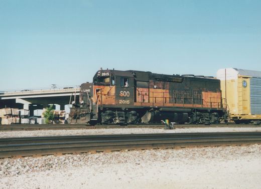 A side view of a Soo Line GP38-2 diesel locomotive numbered 2015 on a railroad main line, pulling a yellow freight car under a clear sky.
