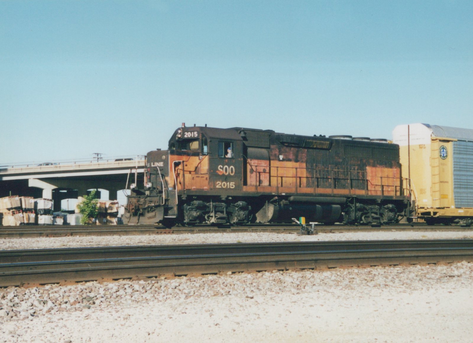 A side view of a Soo Line GP38-2 diesel locomotive numbered 2015 on a railroad main line, pulling a yellow freight car under a clear sky.
