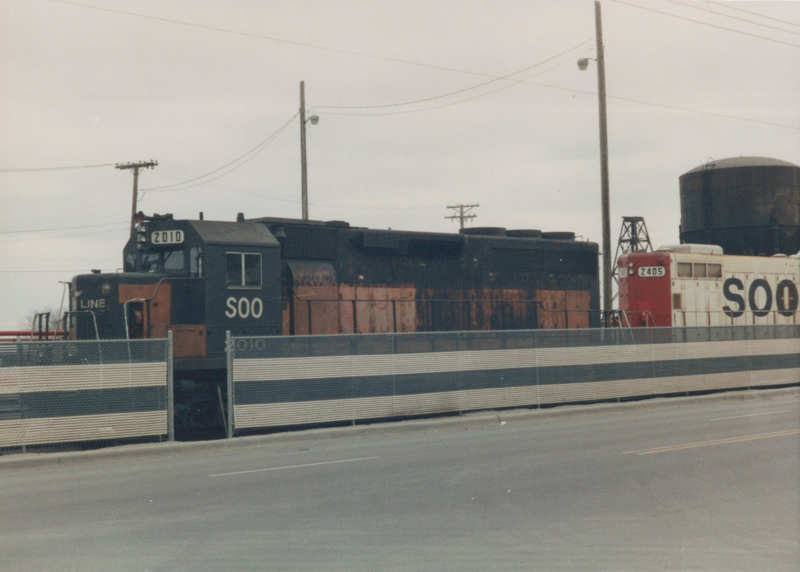 Two Soo Line locomotives, numbered 2010 and 2405, stand side by side on an elevated track behind a striped barrier, with a water tower in the background.