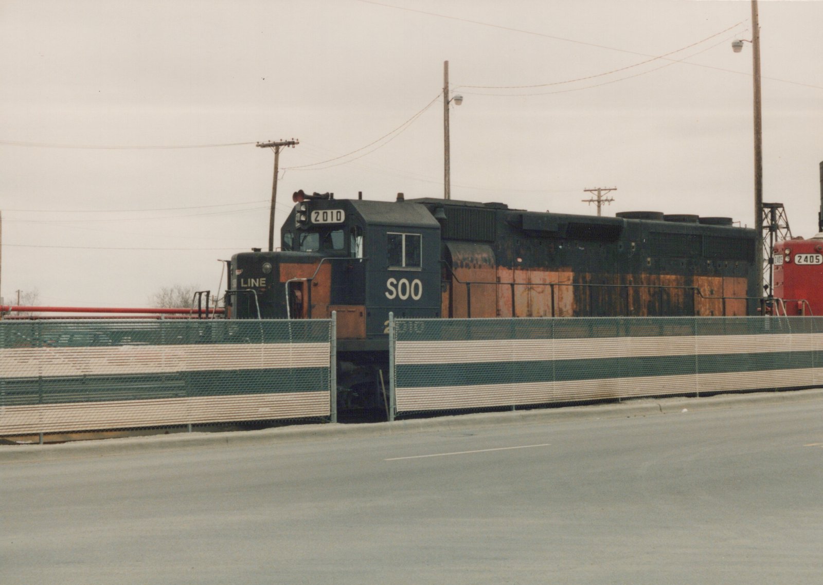 A dark blue and orange SOO Line SD40 locomotive numbered 2010 and a red SOO Line GP38-2 numbered 2405 are parked behind a fence in a rail yard on an overcast day.