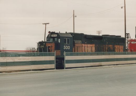 A dark blue and orange SOO Line SD40 locomotive numbered 2010 and a red SOO Line GP38-2 numbered 2405 are parked behind a fence in a rail yard on an overcast day.