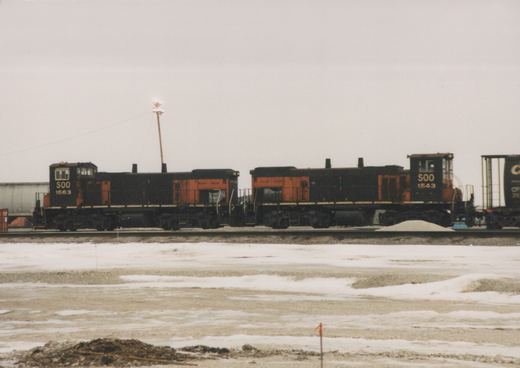 Two black and orange SOO Line diesel locomotives numbered 1563 and 1543 are coupled on a track in a snowy yard, with a freight car behind them.