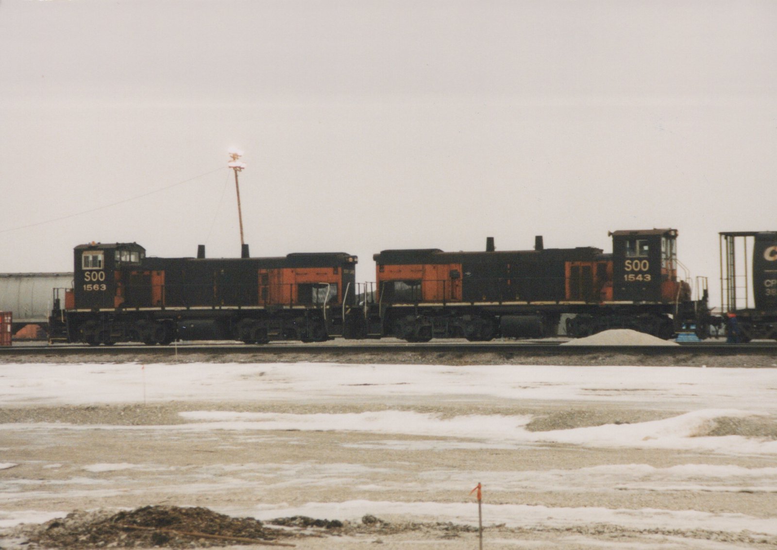 Two black and orange SOO Line diesel locomotives numbered 1563 and 1543 are coupled on a track in a snowy yard, with a freight car behind them.