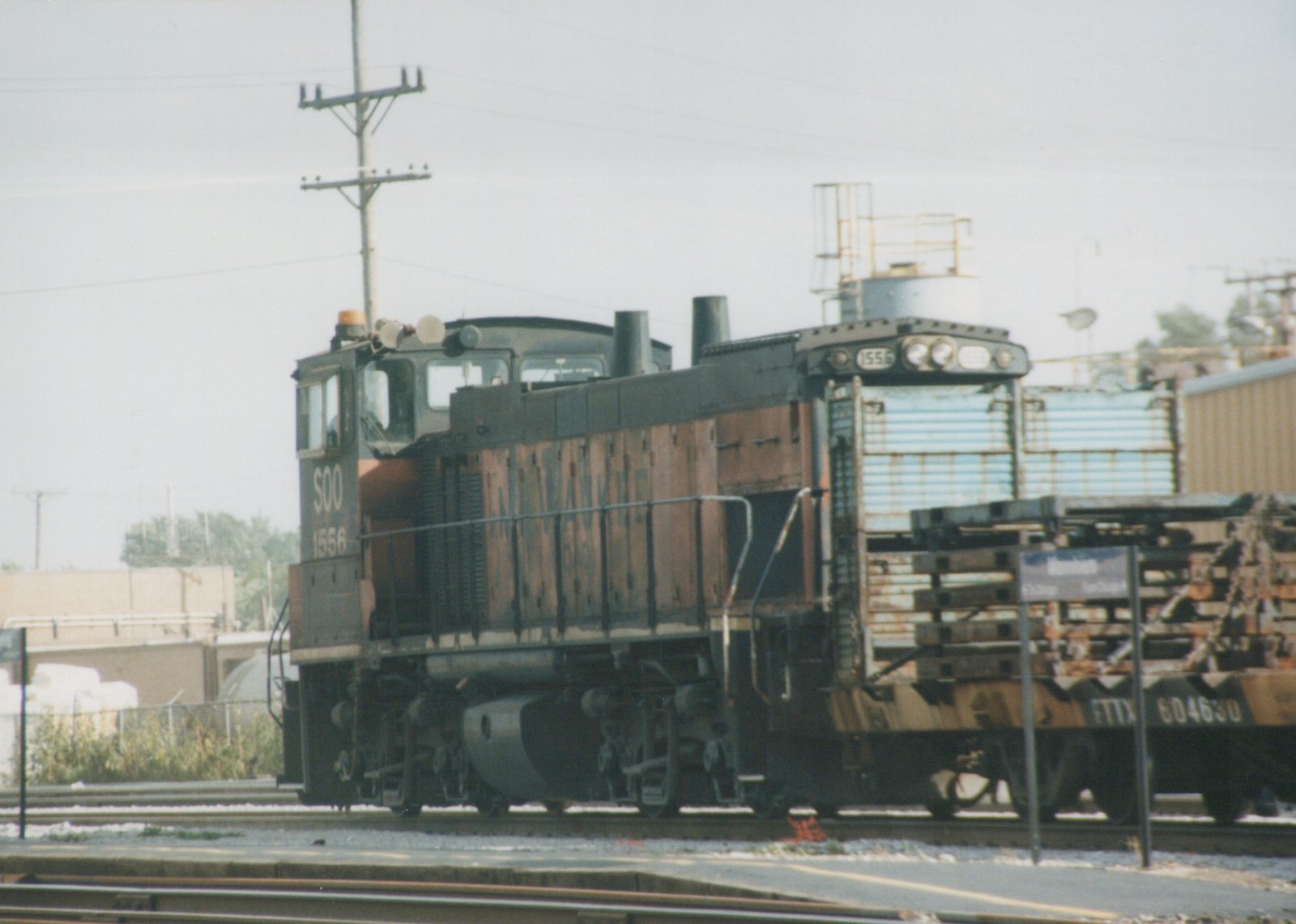 A black and orange SOO Line diesel locomotive numbered 1556 is stopped at a station platform, with a freight car behind it and a sign reading Franklin Park.