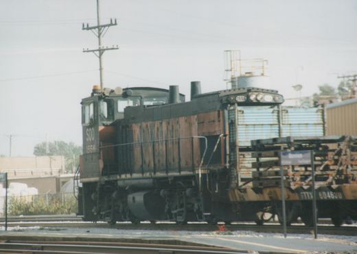 A black and orange SOO Line diesel locomotive numbered 1556 is stopped at a station platform, with a freight car behind it and a sign reading Franklin Park.