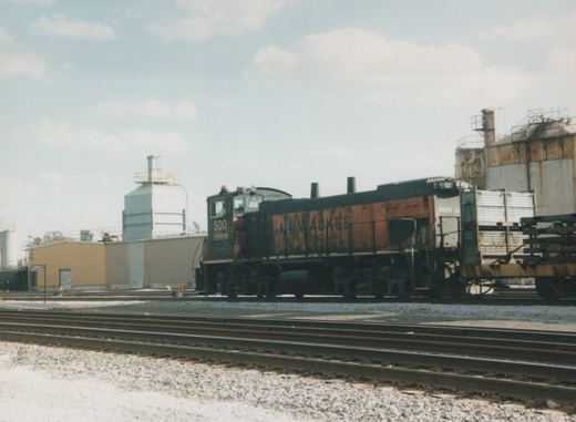 A black and orange SOO Line diesel locomotive number 1556, in a rail yard with multiple tracks, industrial buildings, and a clear sky overhead.