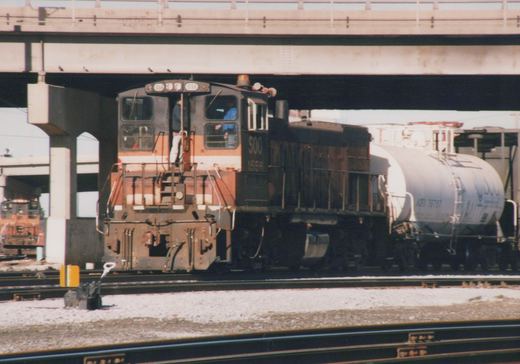 A black and orange Soo Line diesel locomotive numbered 1556 is shown in three-quarter front view, coupled to a white tank car, passing under a concrete overpass on a sunny day.