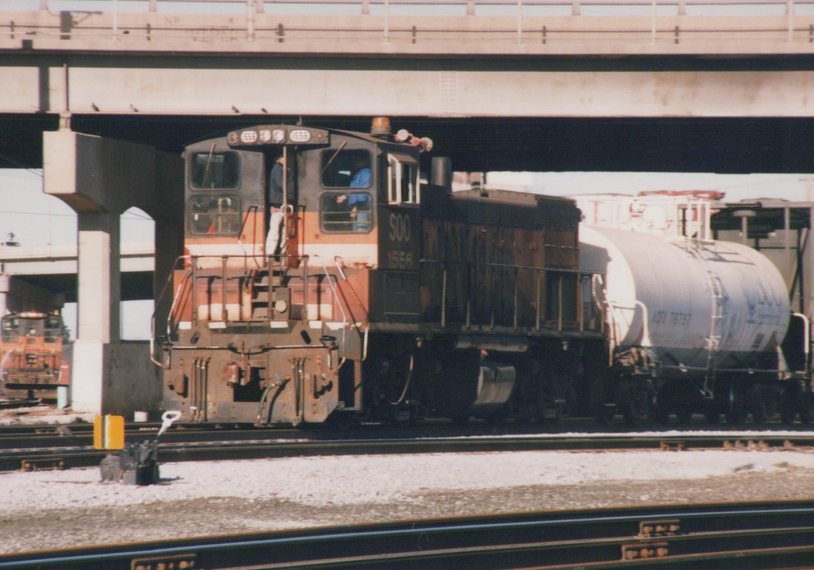 A black and orange Soo Line diesel locomotive numbered 1556 is shown in three-quarter front view, coupled to a white tank car, passing under a concrete overpass on a sunny day.