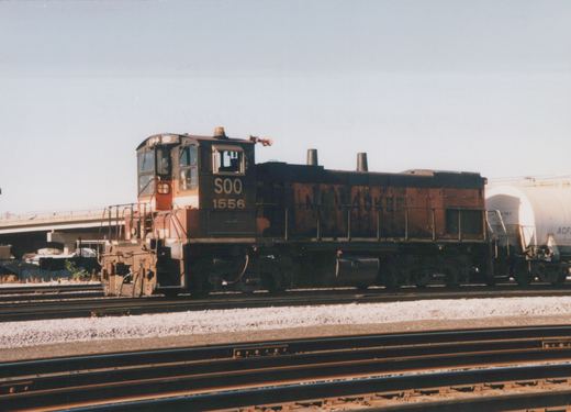 A black, orange, and white diesel locomotive numbered SOO 1556, with faded Milwaukee Road lettering on its side, is on a set of tracks in a rail yard.