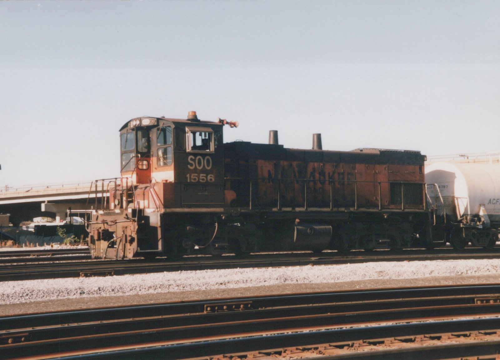 A black, orange, and white diesel locomotive numbered SOO 1556, with faded Milwaukee Road lettering on its side, is on a set of tracks in a rail yard.
