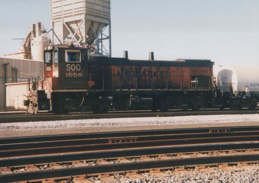 Side view of a Milwaukee Road diesel locomotive number 1556 in a rail yard with gravel ballast and multiple tracks.