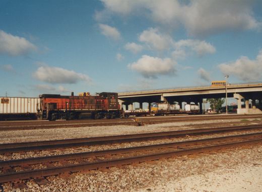 A red and black Soo Line diesel locomotive sits on tracks in a yard under a bridge, with a yellow school bus driving above and freight cars nearby.