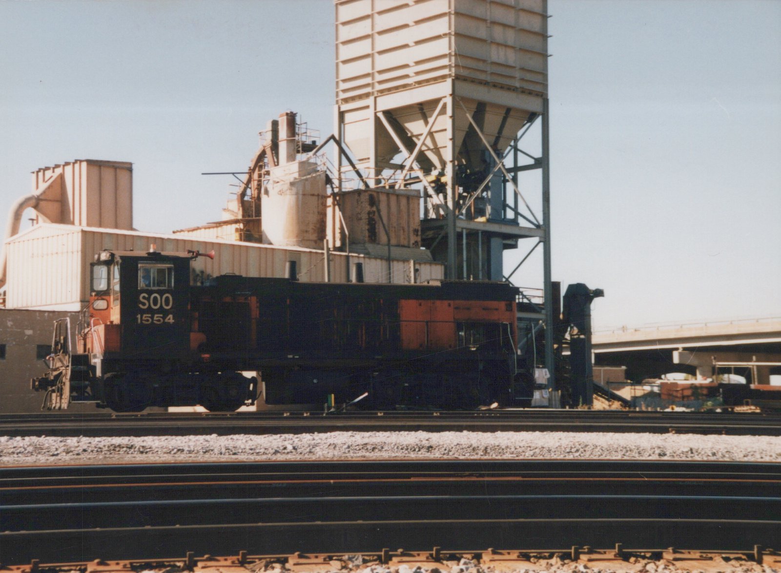 A black and orange Soo Line 1554 diesel locomotive is parked on multiple tracks in a rail yard, with a large industrial structure behind it under a clear sky.