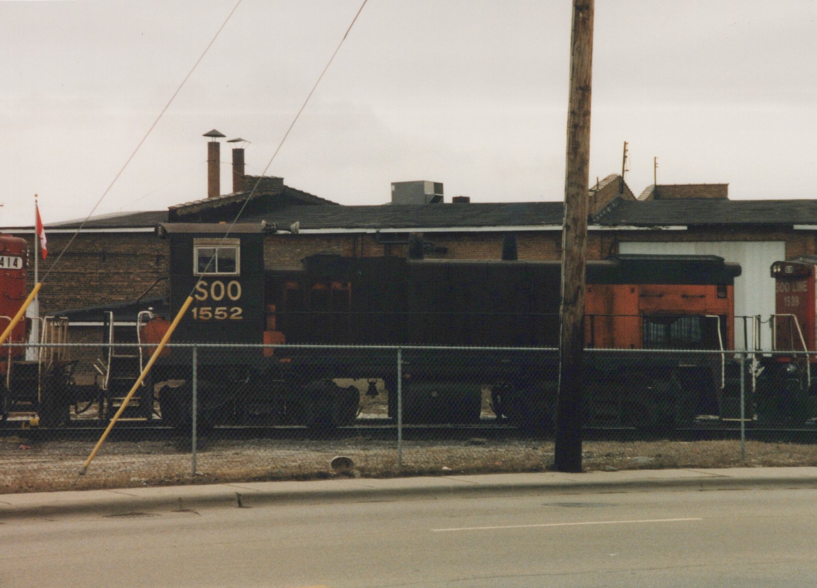 Three Soo Line diesel locomotives parked behind a chain-link fence in a yard on an overcast day.