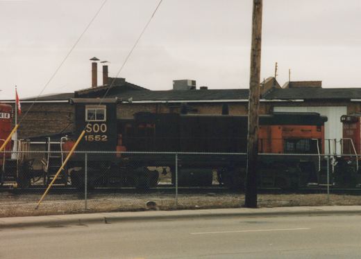 Three Soo Line diesel locomotives parked behind a chain-link fence in a yard on an overcast day.