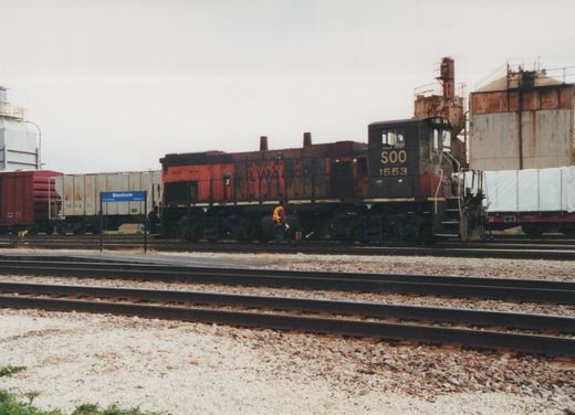 A black and red Soo Line diesel locomotive, number 1553, is stopped at a station with several freight cars in the background. A person in a safety vest walks alongside the tracks. A blue station sign reads 'Mannheim To Chicago Platform 2'.