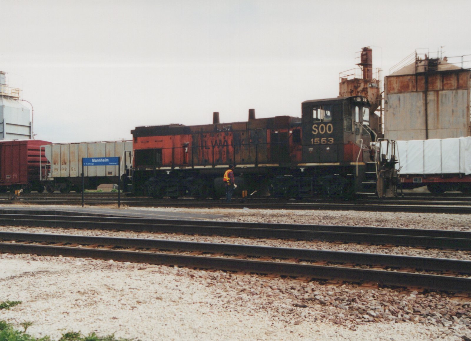 A black and red Soo Line diesel locomotive, number 1553, is stopped at a station with several freight cars in the background. A person in a safety vest walks alongside the tracks. A blue station sign reads 'Mannheim To Chicago Platform 2'.