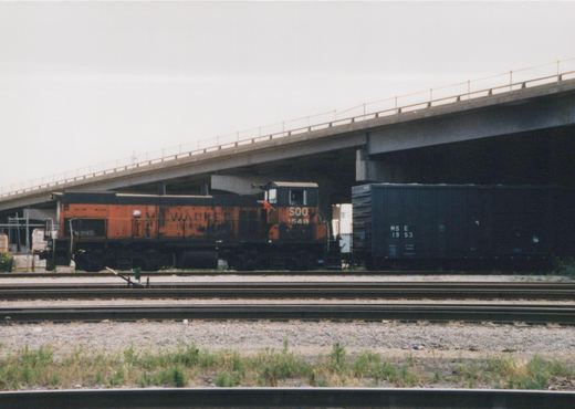 An orange and black SOO Line locomotive numbered 1548 is parked under a concrete bridge next to a dark freight car marked MStE 1953, with multiple rail tracks in the foreground.