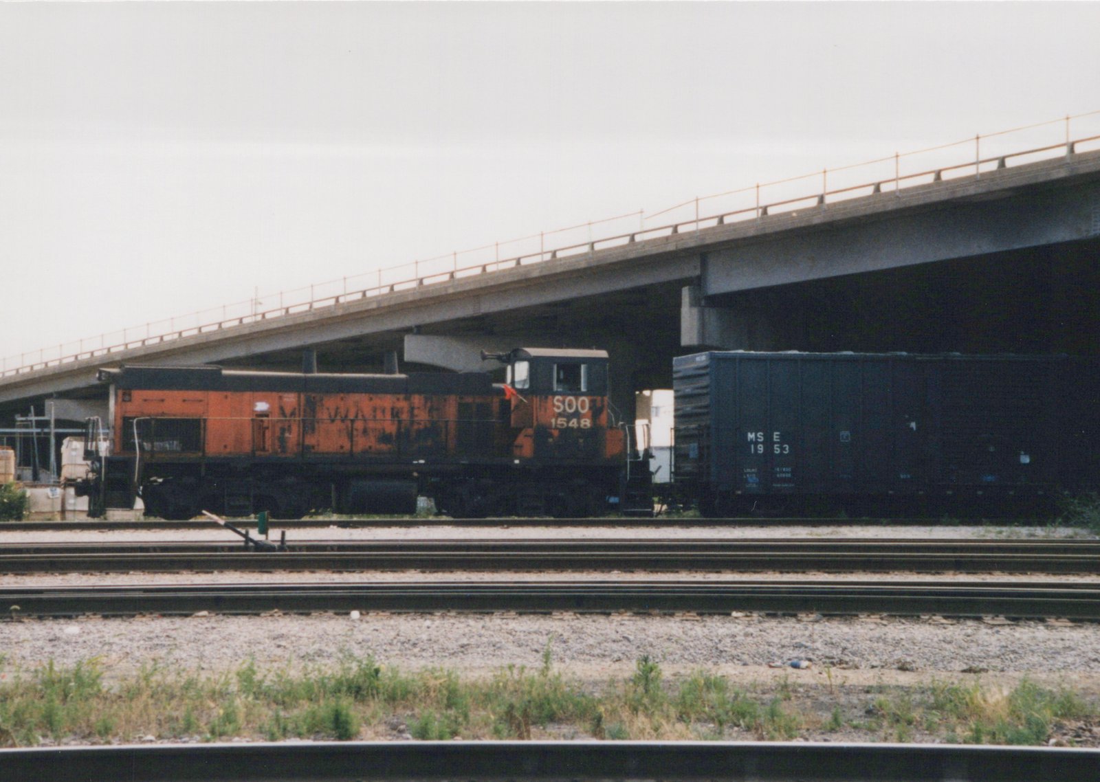 An orange and black SOO Line locomotive numbered 1548 is parked under a concrete bridge next to a dark freight car marked MStE 1953, with multiple rail tracks in the foreground.