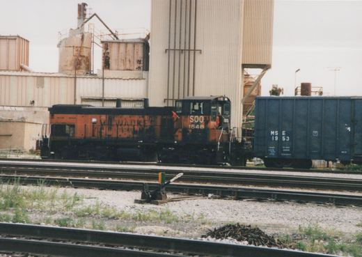 Side view of a Soo Line locomotive numbered 1548, with faded Milwaukee Road lettering, sitting on tracks next to a blue freight car marked MS E 1953, in front of a large industrial silo.