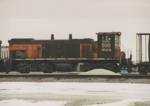 A side view of a black and orange Soo Line diesel locomotive numbered 1543, standing on tracks in a snow-covered yard with a person nearby.