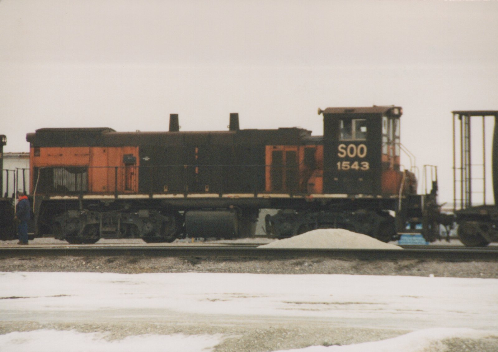 A side view of a black and orange Soo Line diesel locomotive numbered 1543, standing on tracks in a snow-covered yard with a person nearby.