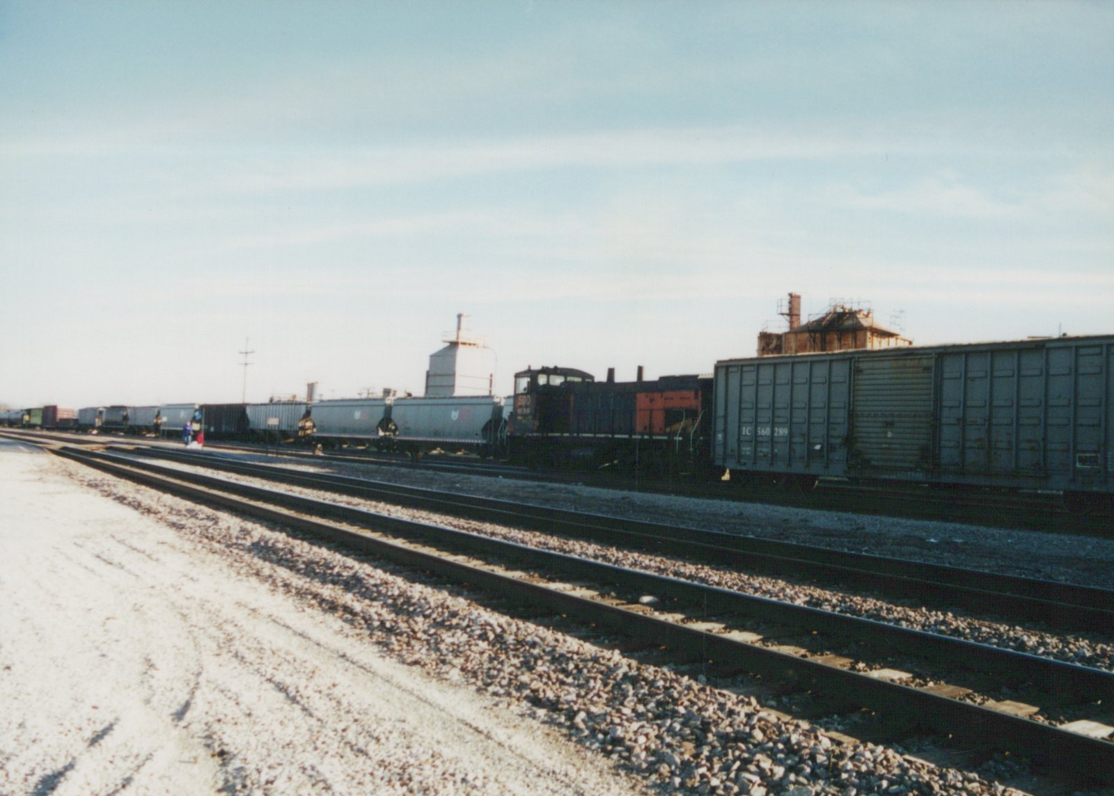 A train with a dark Soo Line locomotive and gray boxcars sits on tracks in a snowy yard under a clear sky.