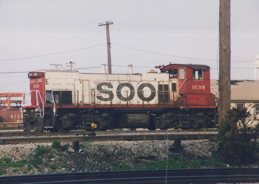 Side view of a red and white SOO Line diesel locomotive number 1539 on railroad tracks, with a water tower and freight cars in the background.