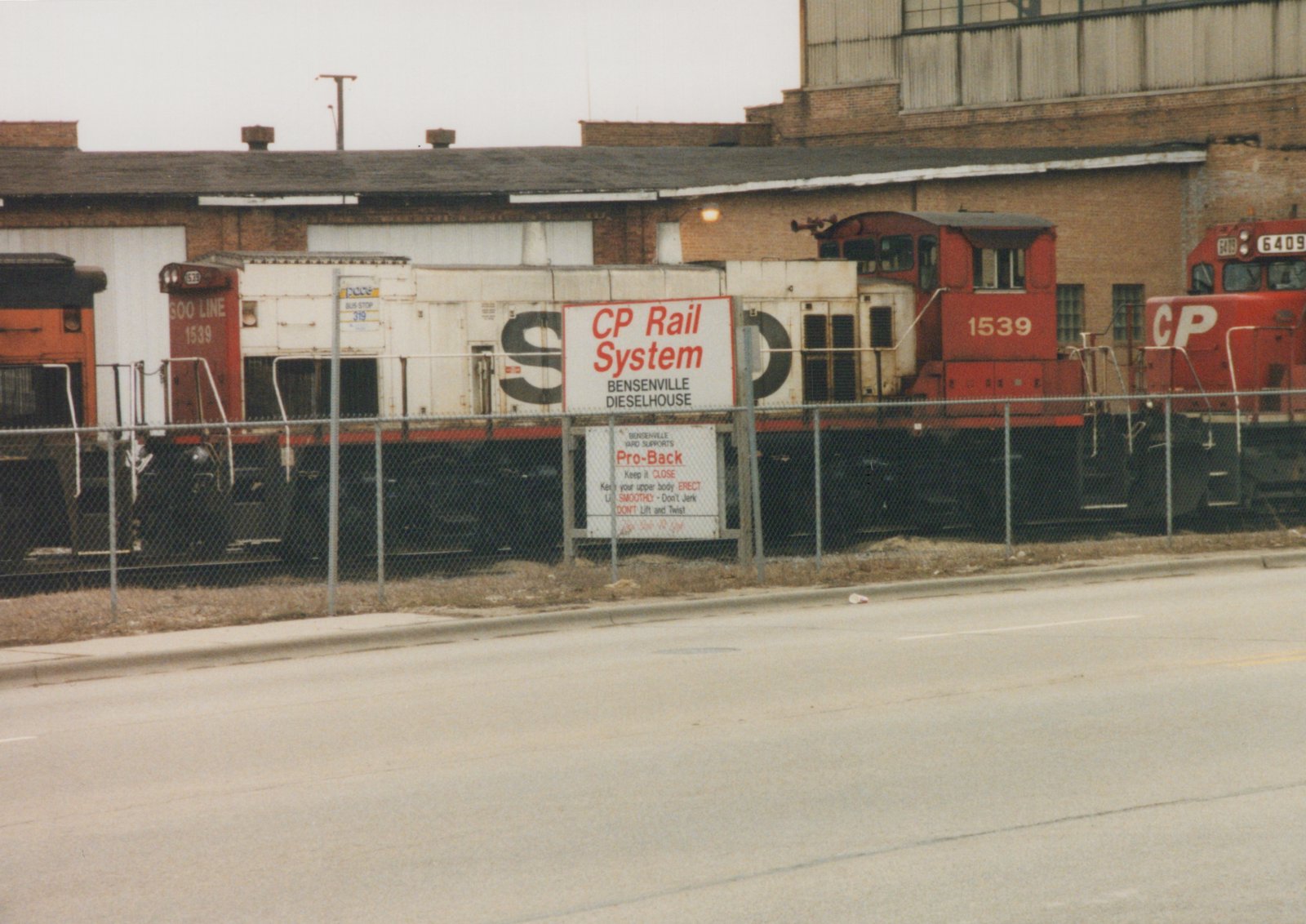 A side view of two red diesel locomotives, one marked SOO LINE 1539 and the other CP 6409, parked behind a chain-link fence in front of a brick dieselhouse building with signs reading CP Rail System and Bensenville Dieselhouse.