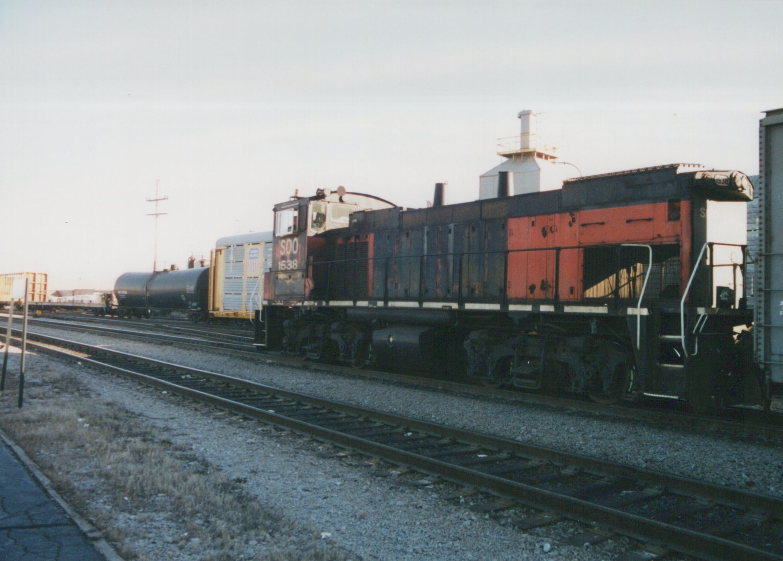 SOO Line diesel locomotive number 1538, painted in black and orange with white trim, is shown on a railroad track pulling freight cars in a yard under an overcast sky.