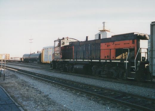 SOO Line diesel locomotive number 1538, painted in black and orange with white trim, is shown on a railroad track pulling freight cars in a yard under an overcast sky.