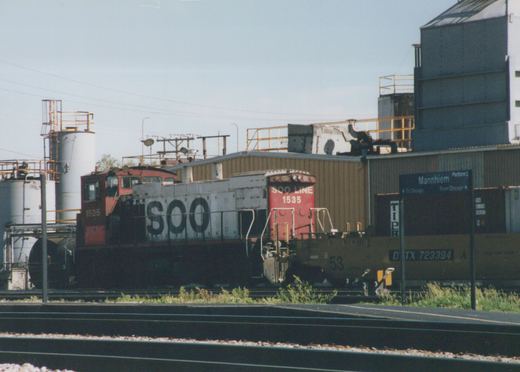 A red and white SOO Line diesel locomotive number 1535 is parked next to a brown freight car in a yard with a station sign for Mannheim Platform 2 visible in the background.