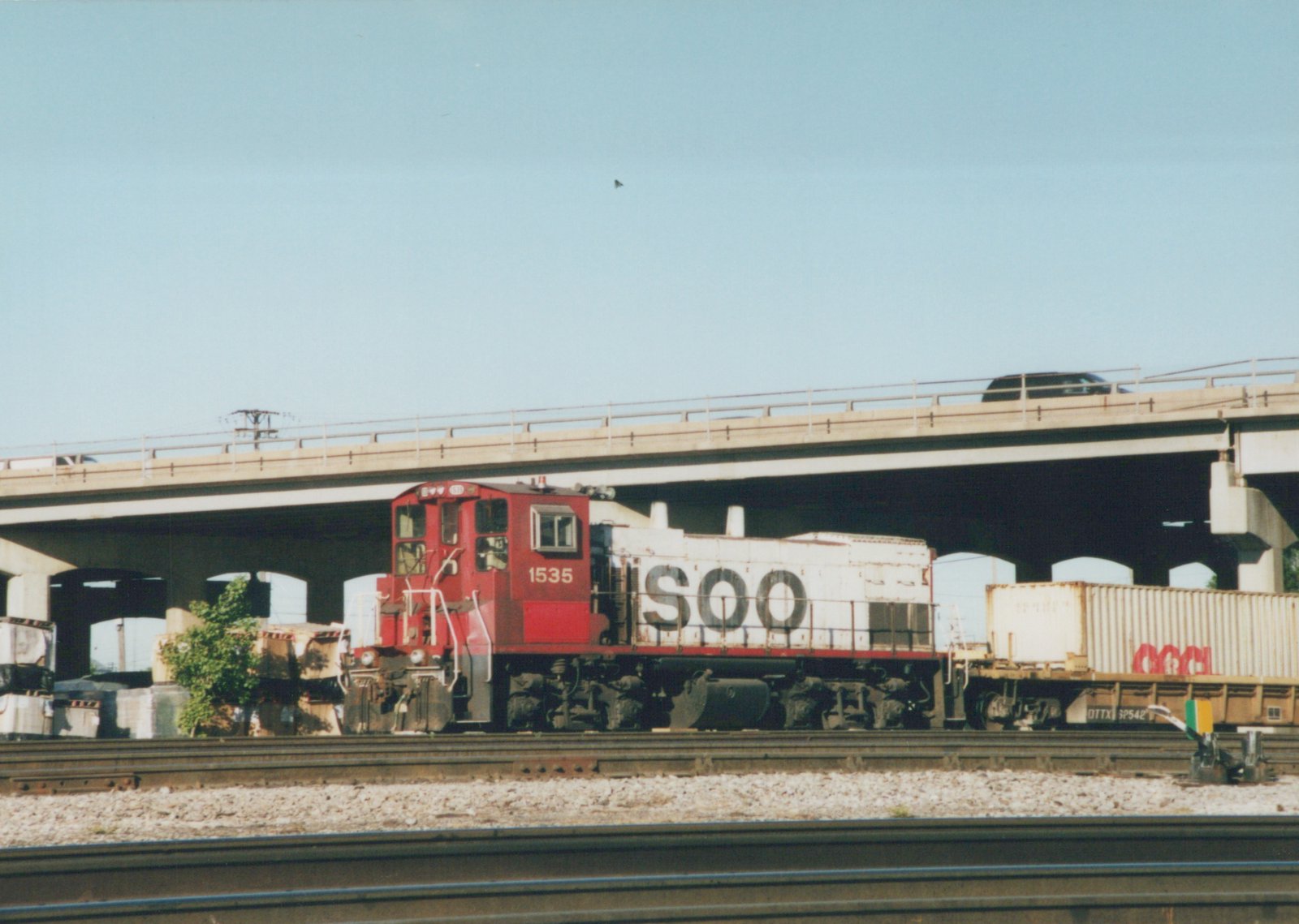 A red and white Soo Line locomotive numbered 1535 is shown from the side, pulling freight cars under a highway overpass on a sunny day.